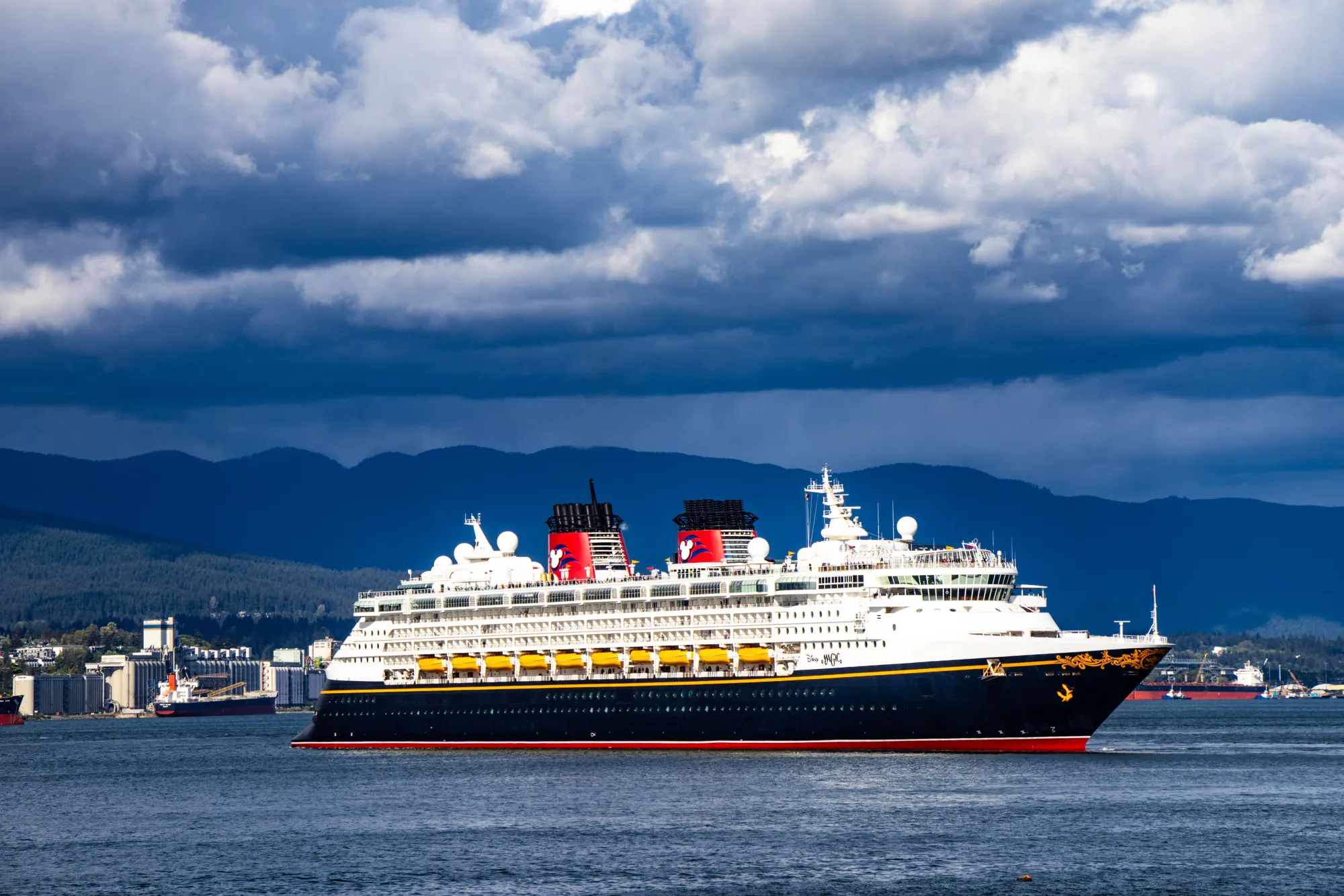 Disney Cruise Line ship sailing past a forested coastline under dramatic clouds